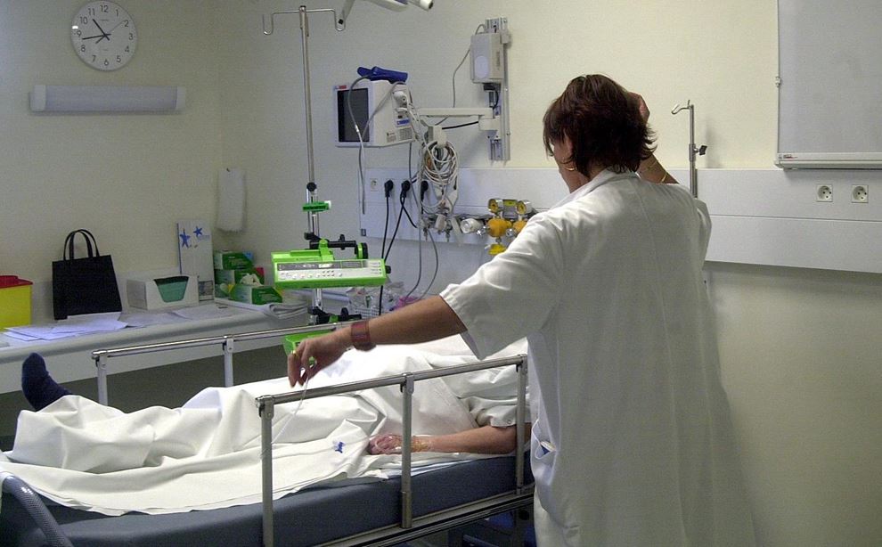 Kvinde i hvid kittel foran hospitalsseng med patient. 